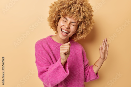 Positive carefree woman with curly hair keeps hand near mouth as if microphone dressed in casual pink jumper sings song isolated over beige background. People happy emotions and fun concept.