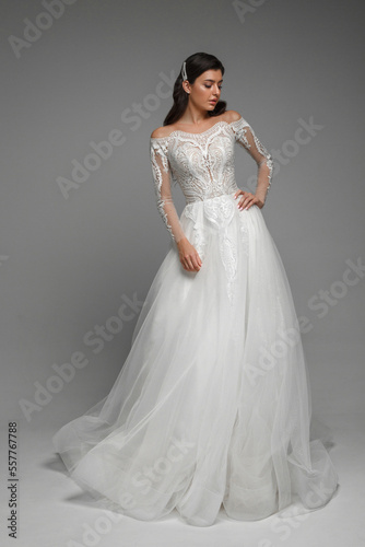 Full lenght of the young brunette bride wearing a new wedding white dress with the patterns in it. Female shotting in the studio, standng and posing on the grey background.