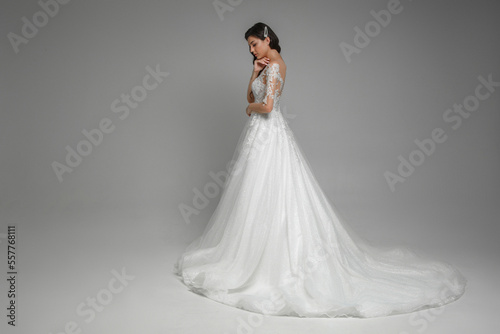 Side view of the beautiful caucasian brunette bride in the beautiful white wedding dress with pearls and embroidery standing  posing with hands in the grey studio.