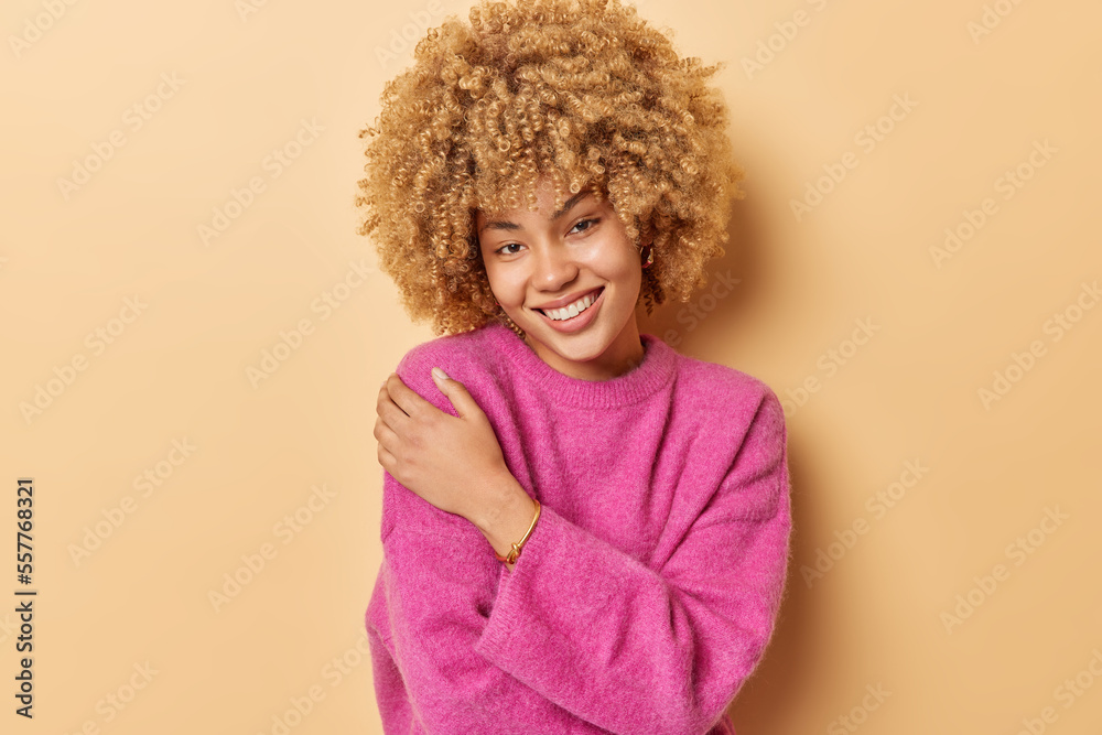 Waist up shot of beautiful curly haired woman embraces herself smiles gently dressed in casual pink jumper expresses self care isolated over brown background. People and pleasant feelings concept Waist up shot of beautiful curly haired woman embraces herself smiles gently dressed in casual pink jumper expresses self care isolated over brown background. People and pleasant feelings concept