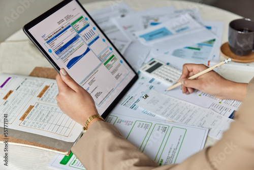 Close up table with utility bills and paper documents. Unknown woman holds tablet writes down notes summarizes cost money uses calculator counts taxes. Bookkeeping and financial paperwork concept