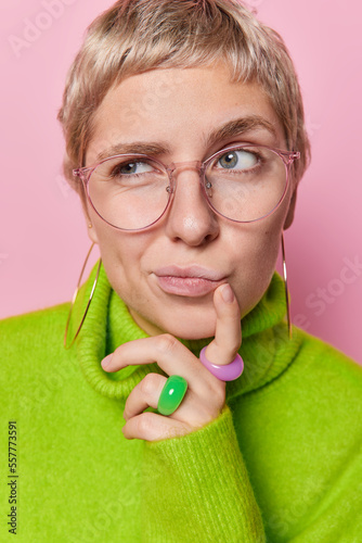 Headshot of thoughtful short haired woman purses lips keeps finger near lips looks pensively aside ponders on something wears big optical eyeglasses and green jumper isolated over pink background