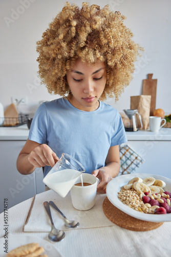 Indoor shot of young curly woman pours milk in coffee going to have breakfaast poses against kitchen interior wears casual blue t shirt being at home during weekend. Tasty cereals with fruits