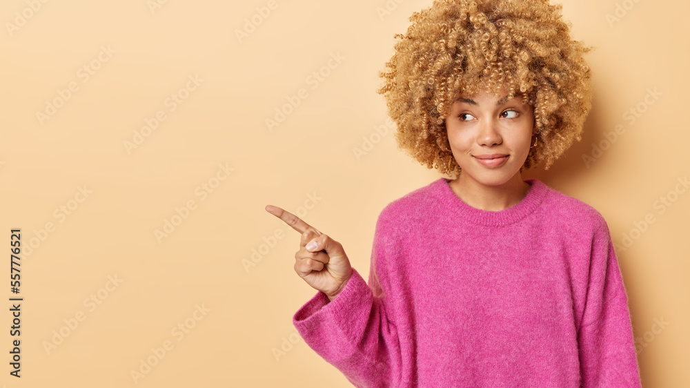 Horizontal shot of good looking woman with blonde curly hair dressed in casual pink jumper points index finger aside shows advertisement shows sale news or promo stands against brown background