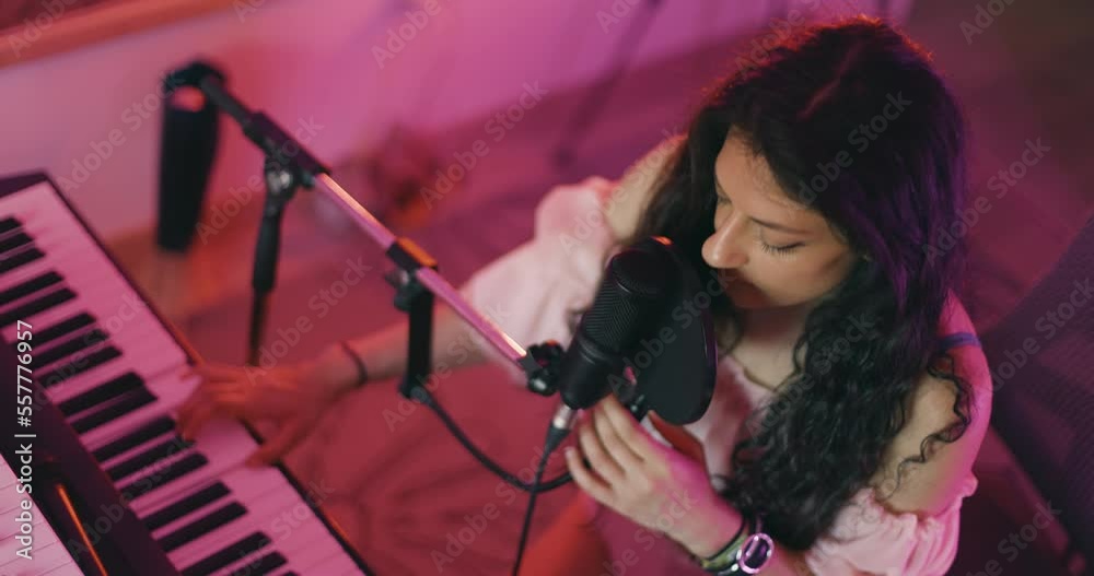 Girl singing on a professional studio microphone and playing on electric keyboard. Female music producer in studio with audio recording equipment. Making a song. Colorful lights.