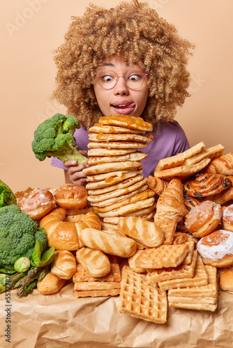 Photo of shocked woman with curly hair licks lips with tongue stares at appetizing sweet dessert holds broccoli surrounded by pile of unhealthy food and green vegetables isolated over beige background