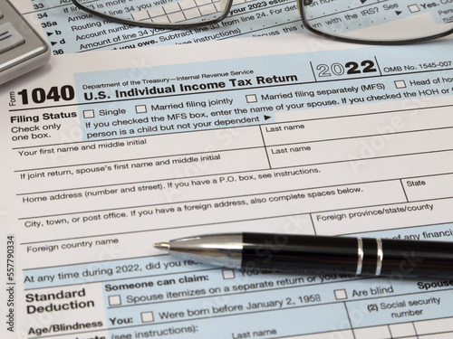 An IRS 1040 tax year 2022 form is shown in 2023, along with an ink pen, calculator, and glasses. The Internal Revenue Service tax filing deadline in 2023 is scheduled for April 18. photo
