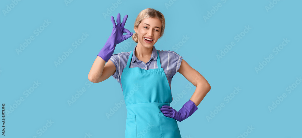 Young woman in apron and rubber gloves showing OK on light blue background Young woman in apron and rubber gloves showing OK on light blue background