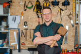 Portrait of a carpenter in goggles and overalls holding a wooden hammer in the workshop against the background of a wall with tools.