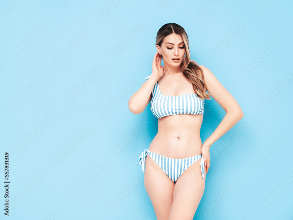 Fototapeta premium Young blond model in summer striped swimwear bathing suit. Sexy carefree woman having fun and going crazy. Female posing near blue wall in studio. Cheerful and happy. Perfect body