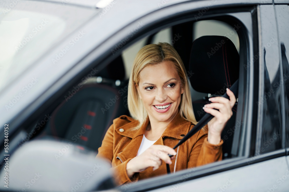 A happy blond woman is putting on seat belt. in her car.