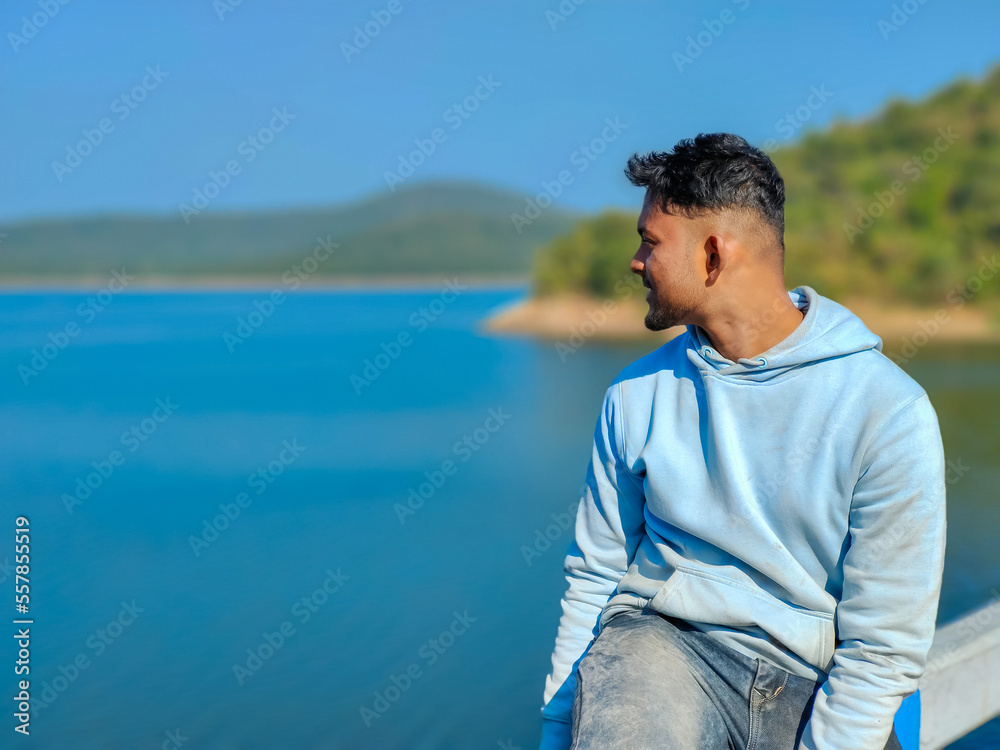 Young man sitting on the bridge and looking at blue river portrait, lake with blue sky scenery. Holiday concept
