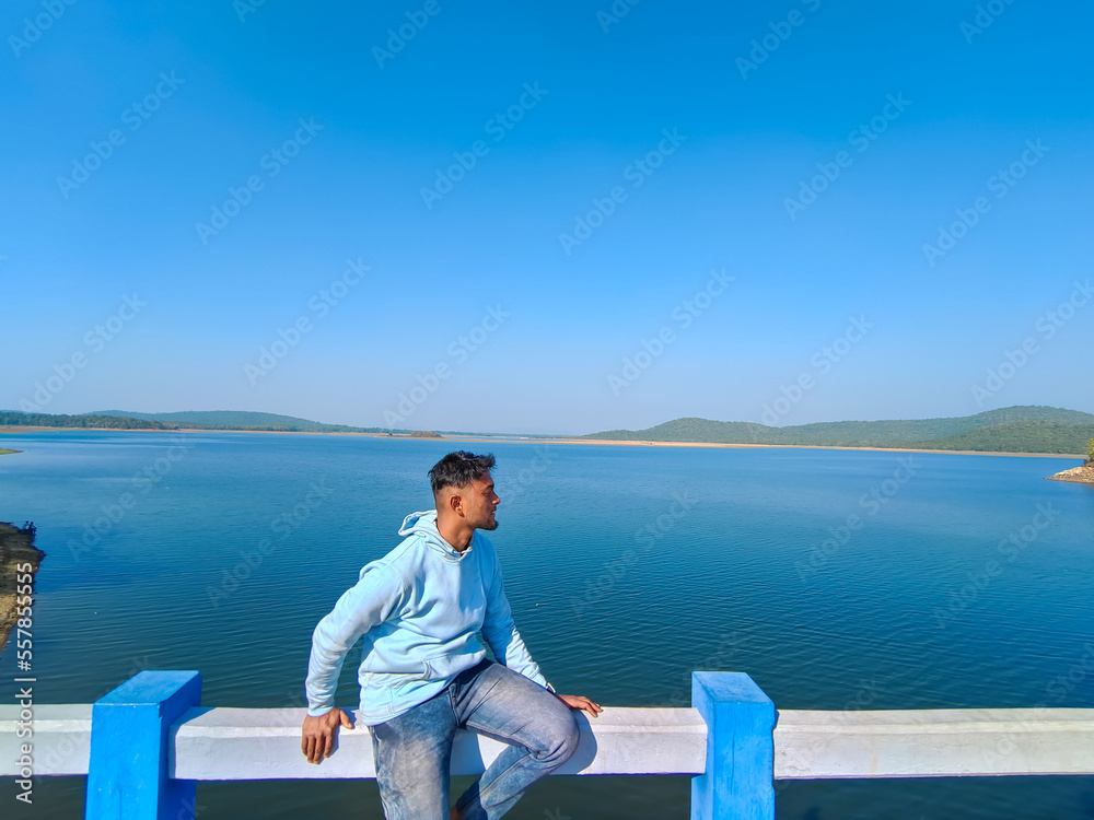 Young man sitting on the bridge and looking at blue river, lake with blue sky scenery. Holiday concept