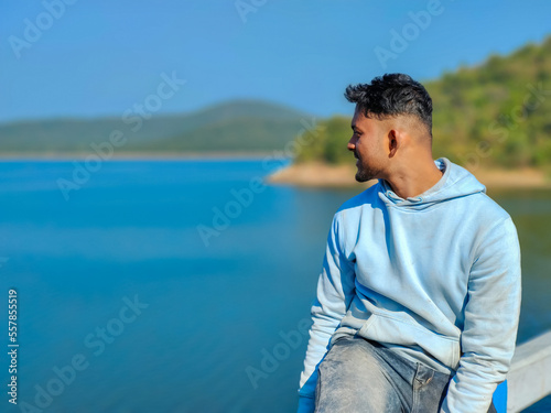 Young man sitting on the bridge and looking at blue river portrait, lake with blue sky scenery. Holiday concept