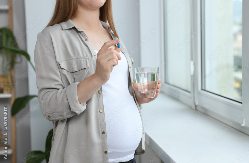 Pregnant woman holding pill and glass with water near window at home, closeup Pregnant woman holding pill and glass with water near window at home, closeup