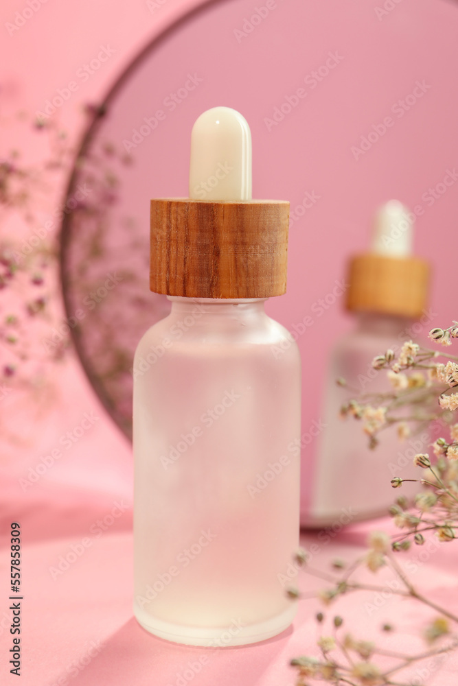 Bottle of face serum and beautiful flowers near mirror on pink background, closeup