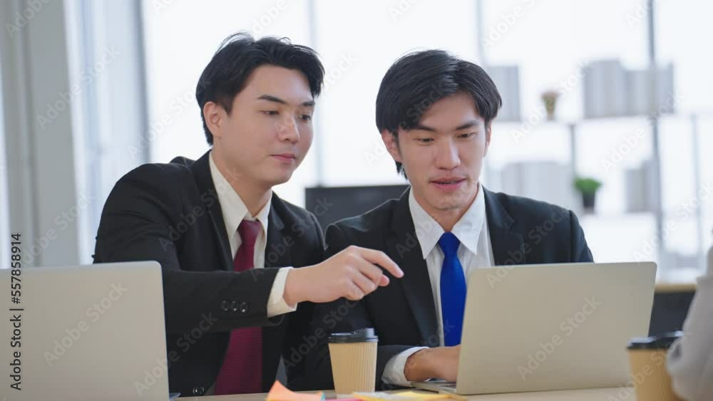 Two young asian businessmen wearing suit discussing financial market data and using laptop at office
