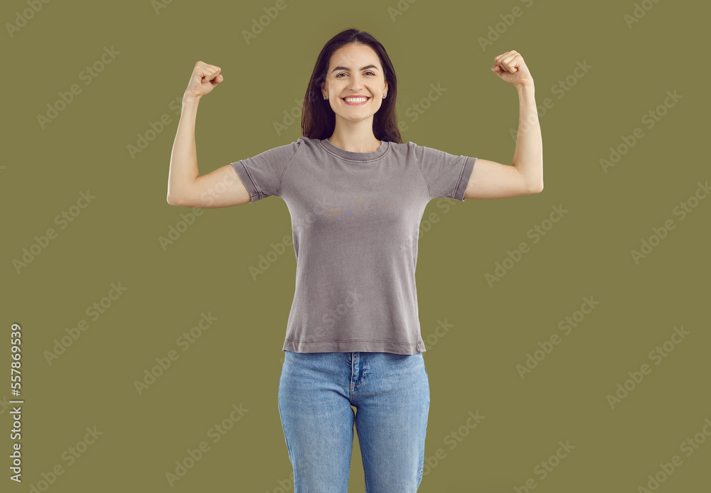Smiling strong powerful young woman showing her biceps. Happy attractive girl wearing jeans and t-shirt raising hands showing muscles. Girl power, feminism concept Smiling strong powerful young woman showing her biceps. Happy attractive girl wearing jeans and t-shirt raising hands showing muscles. Girl power, feminism concept
