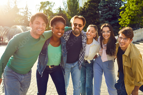 Happy diverse people having fun together. Bunch of six cheerful young Caucasian, Afro American and Asian friends standing in warm evening sunlight, hugging each other, looking at camera and smiling