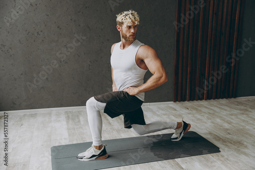 Full body sideways young strong sporty sportsman man wear white tank shirt black shorts leggings do squat lunges akimbo hands on waist at floor warm up training indoor at gym. Workout sport concept. photo