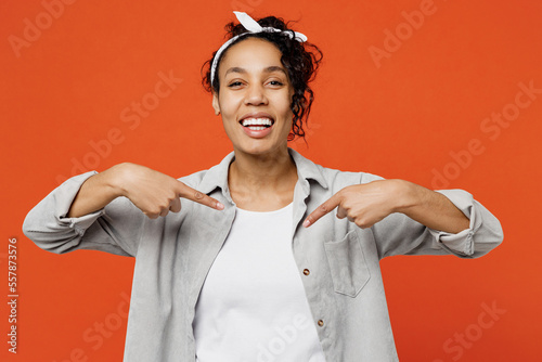 Young smug ambitious fun woman of African American ethnicity she wears grey shirt headband point index fingers on herself isolated on plain orange background studio portrait. People lifestyle concept.