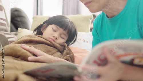 Asian mother hold a book reading story and gentle pat cute little child girl tucked in brown blanket feeling sick, nap and sleep on sofa at home. Parenting love, care, childhood and family lifestyle