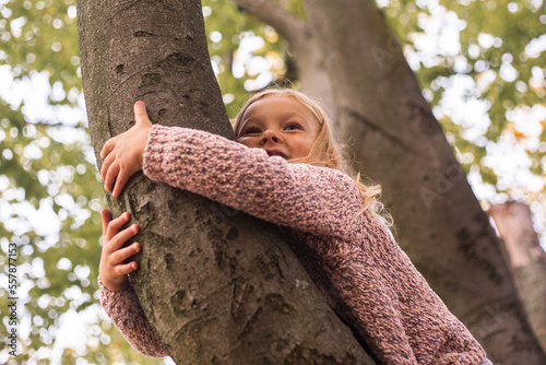 Smiling blond girl child lies hugging a tree with two hands