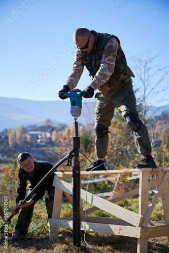 Male workers building pile foundation for wooden frame house. Men builders drilling piles into the ground on blue sky background.