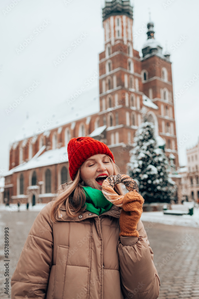 Fototapeta premium Xmas tourist woman in a bright hat and warm jacket holding baked obwarzanek traditional polish cuisine snack bagel on old city Market square in Krakow. Merry Christmas and Happy Holidays. 
