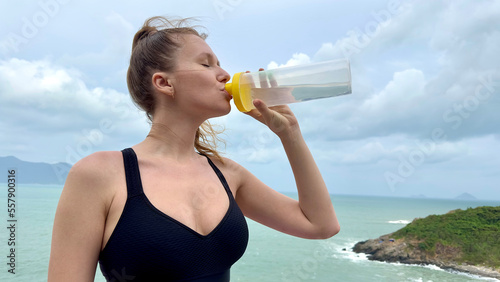 Portrait of happy positive thirsty girl, young beautiful woman is drinking pure fresh mineral water from plastic bottle on natural sea, ocean background in tropical country, holding bottle in hand