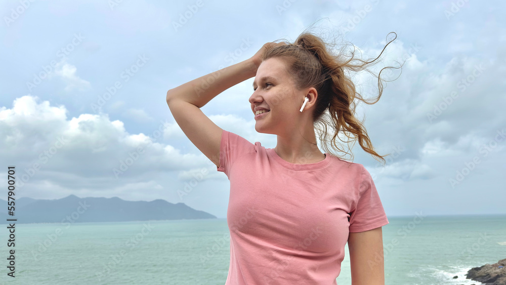 Portrait of beautiful girl, young happy woman at sea, ocean background, enjoying summer sunny day, vacation, have fun, listen to music in wireless earphones on smartphone, using phone on mountain