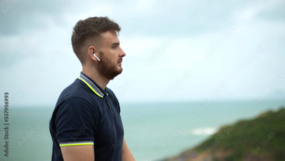 Side portrait of bearded handsome serious pensive thoughtful guy, young man on natural sea, ocean background listening to music, enjoying song in wireless earphones, thinking, dreaming