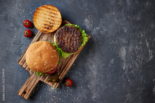 Tasty homemade grilled burger with beef, cheese, tomatoes, pickles, onion and lettuce on a dark concrete background with copy space. Top view flat lay. Fast food and junk food.