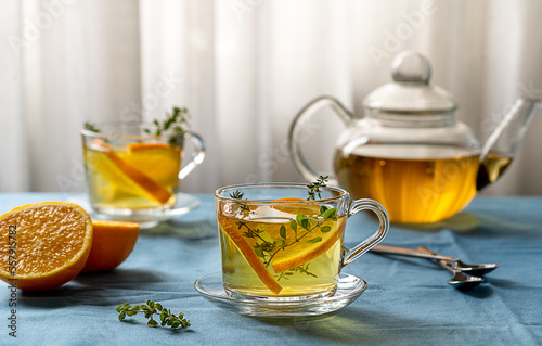 Herbal tea with oregano and thyme and slices of orange in glass cups and teapot with blue background