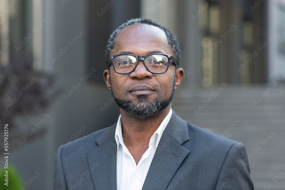 Close up portrait of serious businessman, afro american man in business suit and glasses looking at camera thinking, boss outside office building. Close up portrait of serious businessman, afro american man in business suit and glasses looking at camera thinking, boss outside office building.