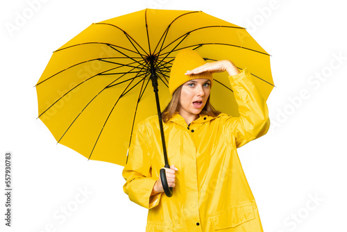 Young blonde woman with rainproof coat and umbrella over isolated chroma key background doing surprise gesture while looking to the side