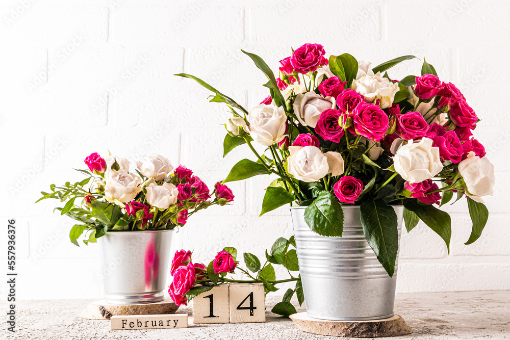 many white and red roses in two metal decorative buckets on a marble table against a white brick wall. wooden calendar with a valentine's day holiday.
