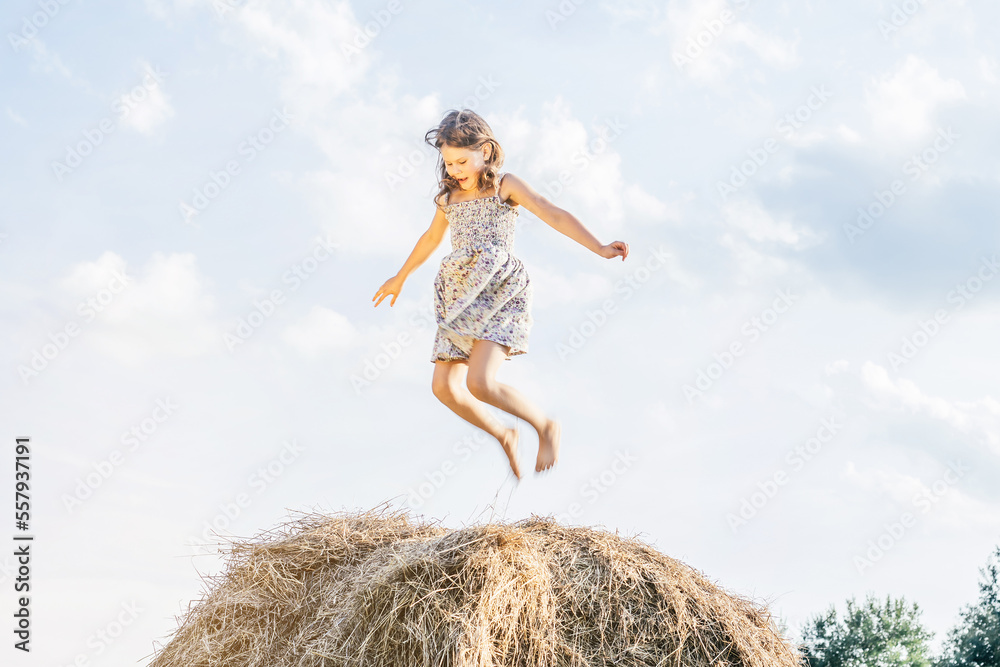 Happy little girl jump up at top of haystack with hands to sides and look down. Have fun on hayrick. Beautiful blue sky.