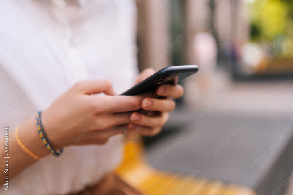 Close-up cropped shot of unrecognizable young woman wearing white jacket holding smartphone and typing online message with both hands on city street. Unknown female holding cellphone in hands outside.