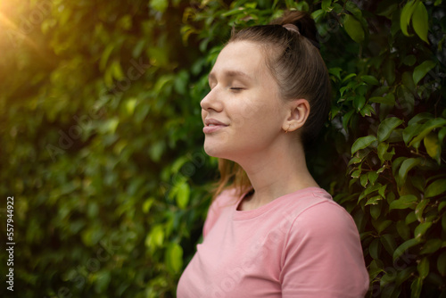 Portrait of a beautiful young woman standing on an Ivy wall or in greenery  smiling and looking away against from plant  girl relaxing outdoors during a sunny summer day. Love for nature  eco life