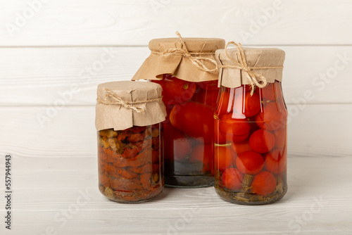 Pickled red cherry tomatoes in a glass jar on a white textured rustic wood. Food preservation for autumn or winter. Home storage. Canning, fermentation concept. Place for text. Copy space.