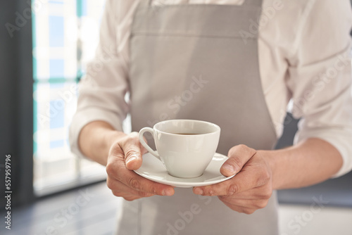 Hands of the waiter in a gray apron holds out a cup of coffee. Barista gives a cup of hot coffee in a cafe, against the background of a large window. High quality photo