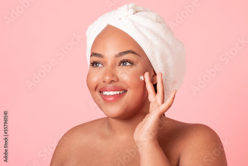 Skin care concept. Closeup portrait of black plus size lady applying moisturizing cream on face, pink background