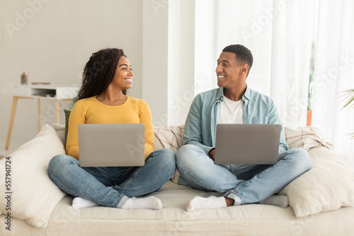 Smiling millennial african american male and female with laptops looking at each other in living room interior