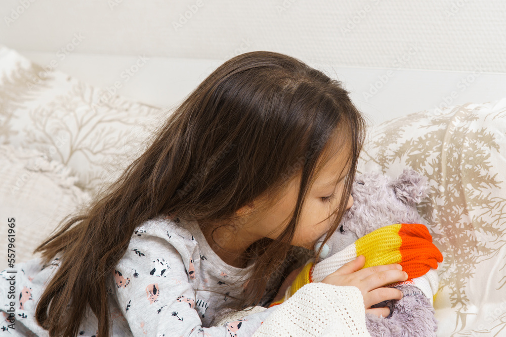 Little girl tie scarf around neck and measure temperature by kissing forehead of sick teddy bear. Kid in sleepwear sitting on bed and playing with stuffed toy. Fever heat, doctor, treatment. Little girl tie scarf around neck and measure temperature by kissing forehead of sick teddy bear. Kid in sleepwear sitting on bed and playing with stuffed toy. Fever heat, doctor, treatment.