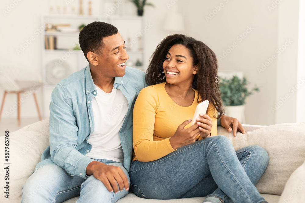 Cheerful millennial african american wife showing smartphone to husband, recommend new app