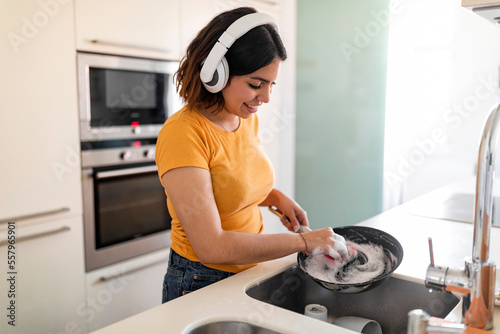Fotografie Arab Woman Washing Pan While Doing Dishes In Kitchen And Listening Music
