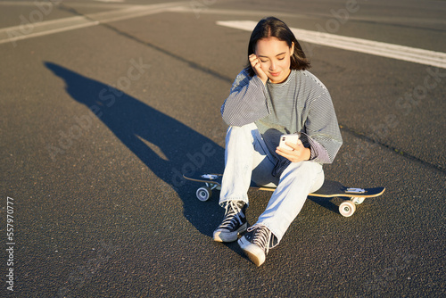 Portrait of asian woman sitting on skateboard, skating on her cruiser longboard, using smartphone app