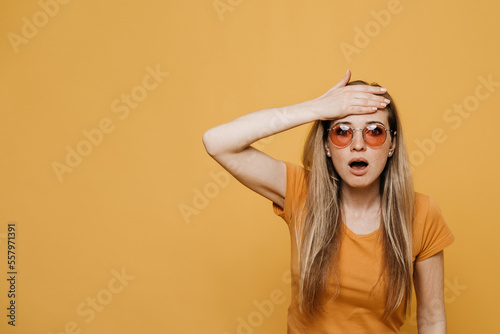 Shocked redhead young woman in red sunglasses, dressed in casual orange t-shirt, touching head in surprise with open mouth, over yellow background. Mock up copy space. Sales and discount.