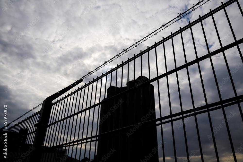 Barbed wire and metal fence with silhouette of tall tower prison factory secret base access and dramatic sky clouds